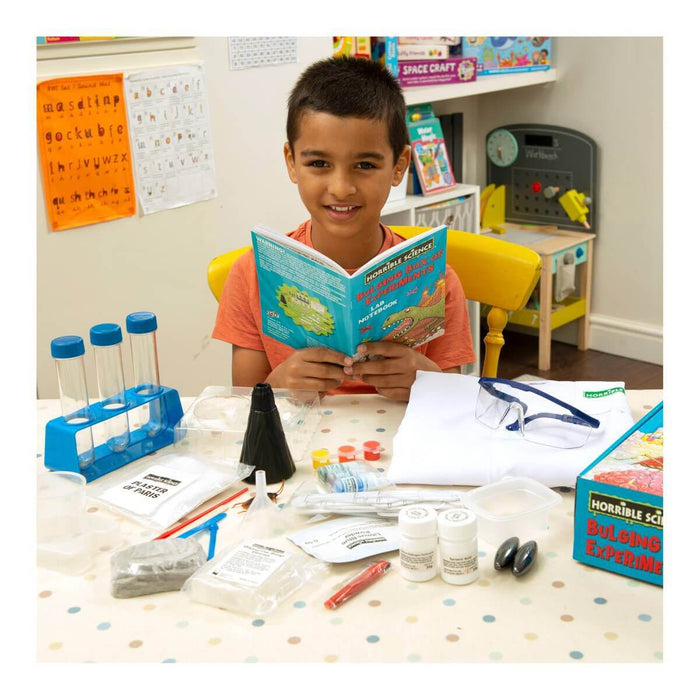 Boy reading a lab notebook at a table with test tubes, safety goggles, a lab coat, and science experiment materials.
