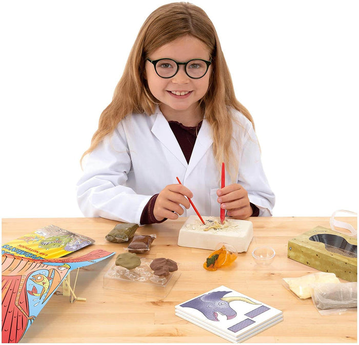 Girl in white lab coat excavating a fossil block with dinosaur models, clay, and science cards on a wooden table.