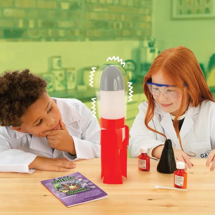 Two children in lab coats watch a red and white toy rocket with science kit bottles and an experiment booklet.