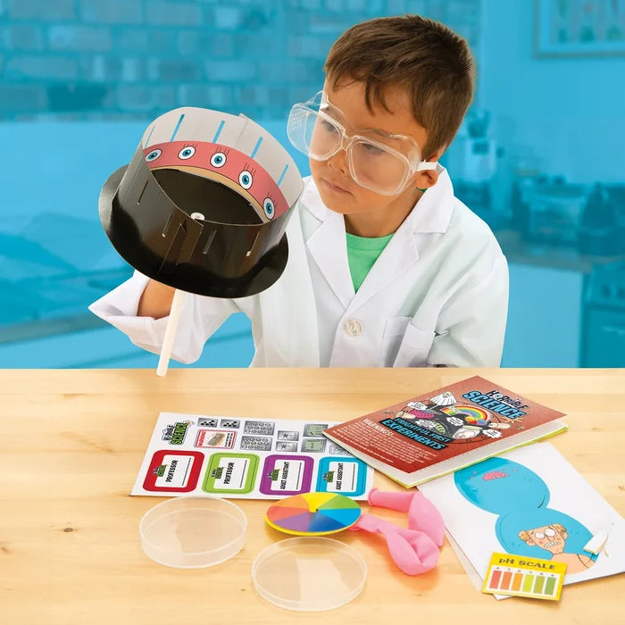 Child in a lab coat and goggles using a science kit with a zoetrope, petri dishes, and balloons on a wooden table.