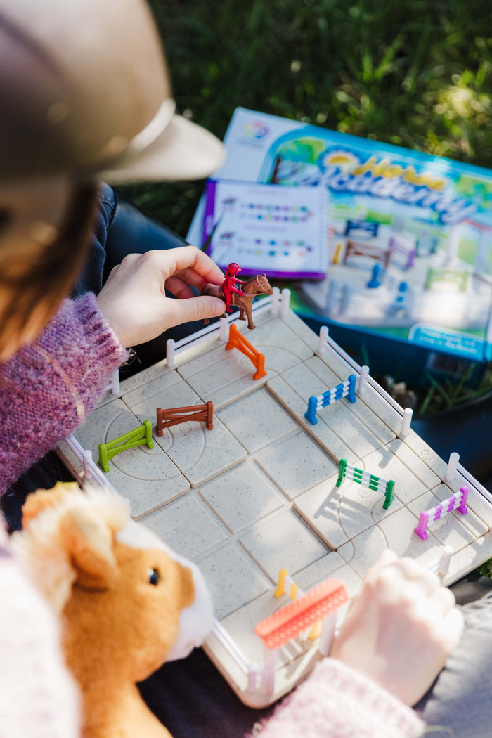 Child playing with a horse jumping puzzle game featuring a grid board, colorful hurdles, and a plastic horse and rider.