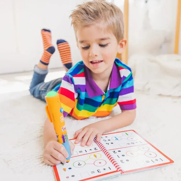 A boy in a rainbow shirt uses a chunky orange interactive pen to solve math problems in a spiral activity book.