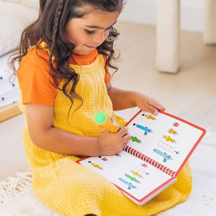 A girl uses a glowing orange pen on a spiral-bound Numberblocks activity book with colorful math puzzles.