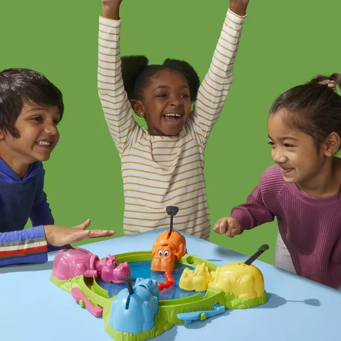 Three children cheer while playing with pink, blue, yellow, and orange hippos on a blue tabletop game base.