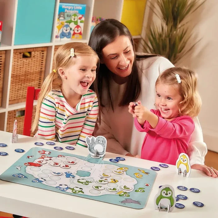 Woman and two young girls playing a penguin board game with blue fish tokens and colorful stand-up characters.