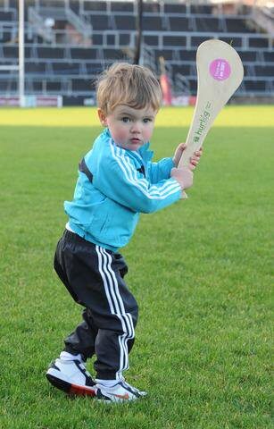 A toddler holding a light-colored Hurlóg soft hurley with a pink logo on a green sports field.