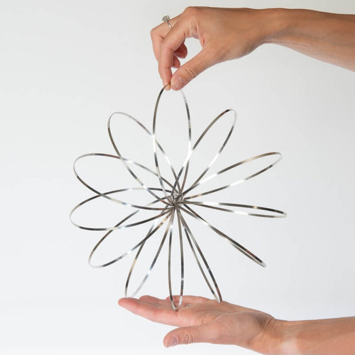 Silver kinetic toy with interlocking metal rings forming a 3D geometric sphere, held between two hands.
