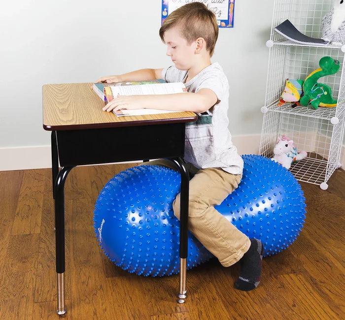 Boy sitting at a desk on a large blue inflatable peanut-shaped sensory roller with small tactile nubs.