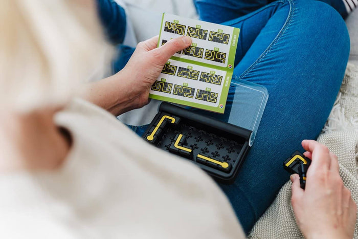Hands holding a green challenge booklet over a black puzzle board with yellow path pieces on blue jeans.