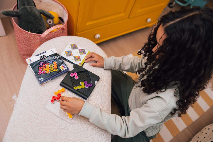 Child plays IQ Gears logic game using colorful gear pieces on a black game board next to its packaging.