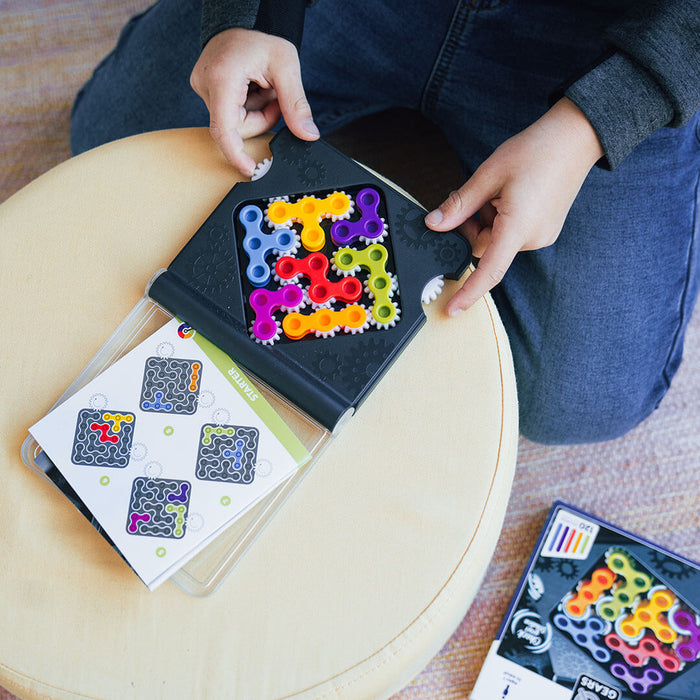 Hands holding a black board with colorful, interlocking gears, showing a challenge booklet and game packaging.