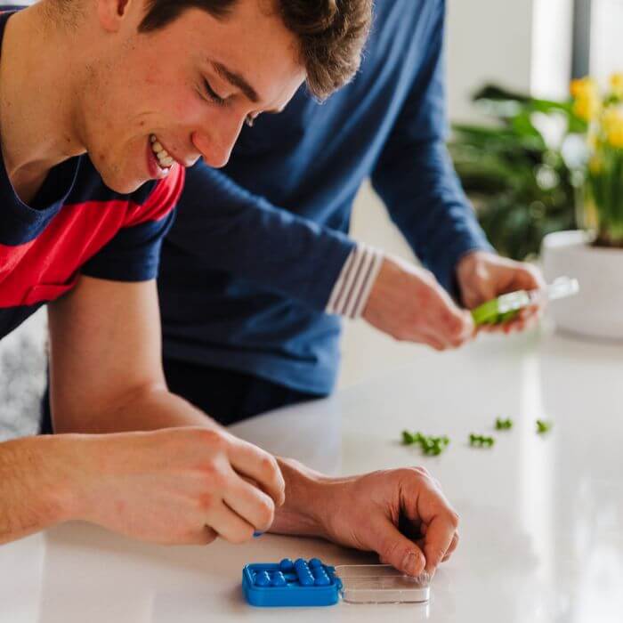 A man smiles while playing with a small blue square puzzle, using his fingers to move tiny blue pegs in its case.
