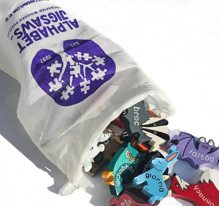 Colorful wooden animal puzzle pieces with Irish names spilling out of a white Alphabet Jigsaws drawstring bag.