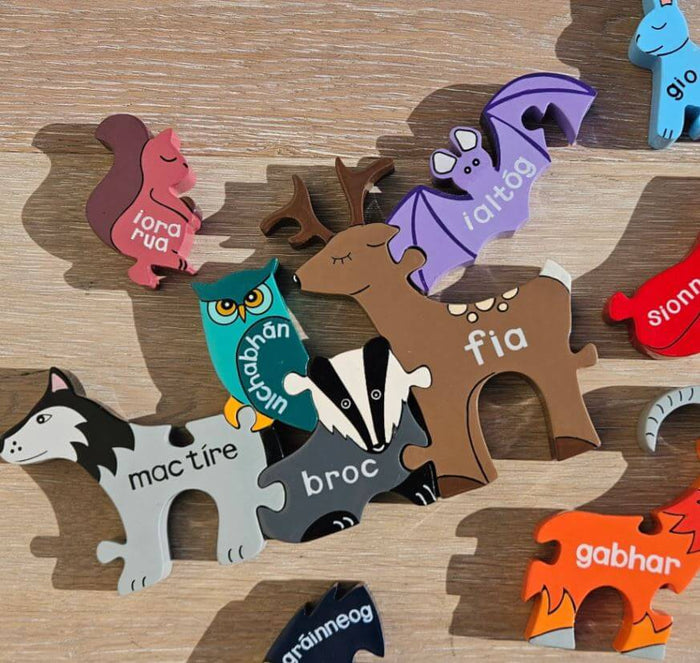 Colorful wooden jigsaw pieces shaped like Irish animals with their names in Irish printed on a light wood surface.