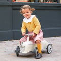 Young girl riding an ivory vintage-style toy car with a chrome grille and silver steering bar on stone pavement.