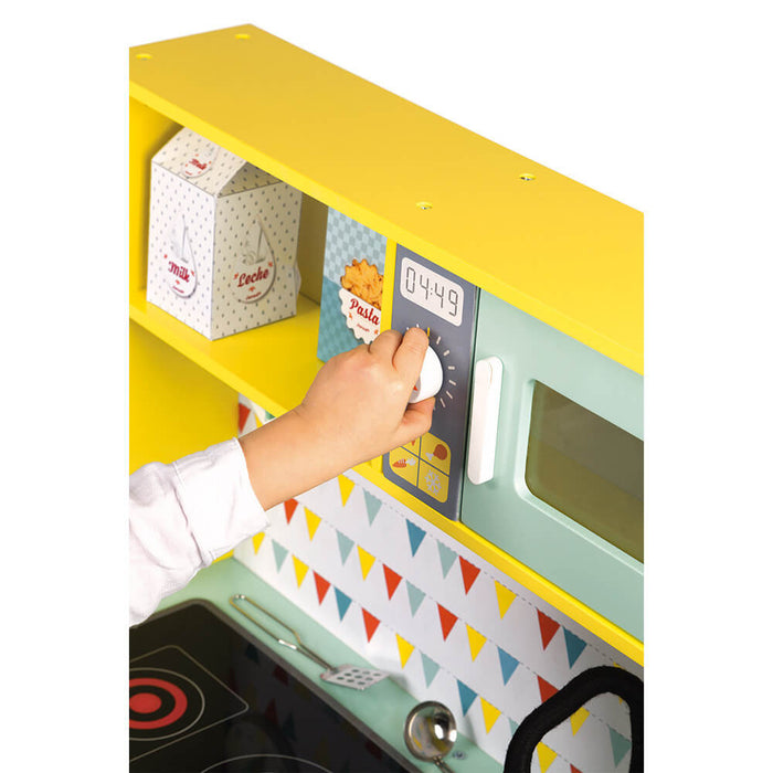 Child's hand turns a white knob on a blue toy microwave in a yellow wooden kitchen with toy food boxes.