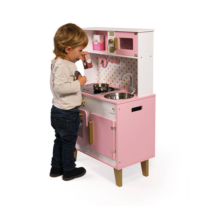 Toddler playing with a pink and white wooden toy kitchen featuring a stove, sink, microwave, and gold accents.