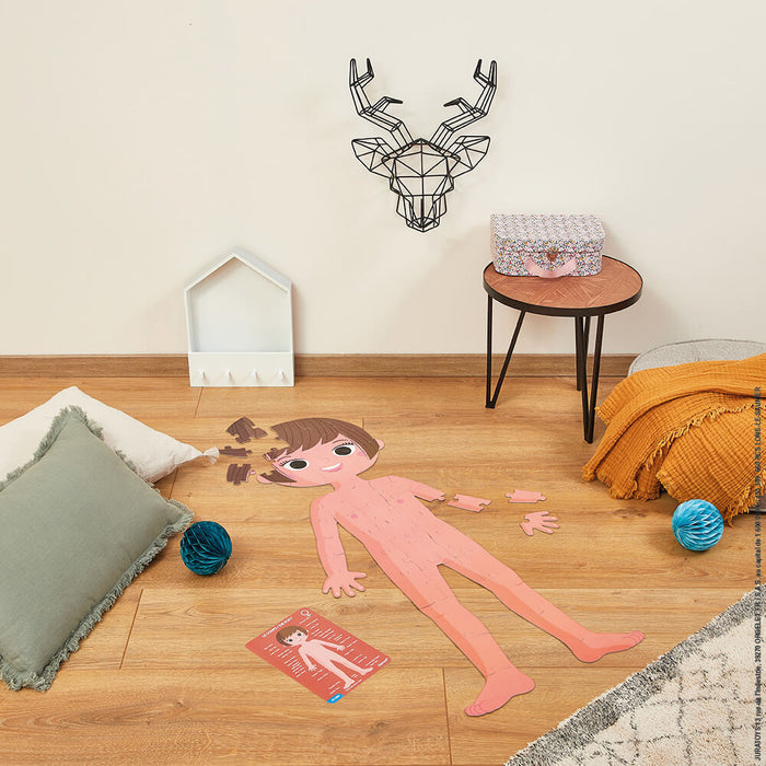 Large pink cartoon floor puzzle of a human body with detached pieces and a reference card on a wooden floor.