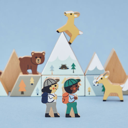 Wooden toy blocks shaped like mountains and forest animals with two hiker figurines on a light blue background.