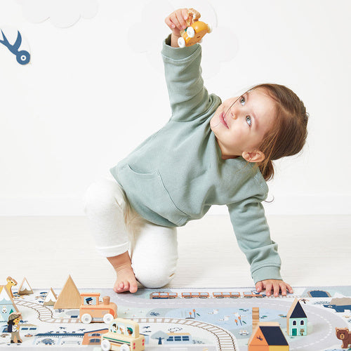Toddler in a green hoodie playing with wooden cars, trains, and mountain blocks on a soft-colored illustrated play mat.
