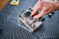 Hand adjusting a knob on a transparent acrylic DJ mixer with a screen and black controls on a gridded workspace.
