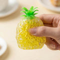 Hand squeezing a squishy yellow pineapple toy filled with small jelly beads, featuring a green leafy top.