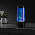 Cylindrical black lamp with blue LED light and two glowing artificial jellyfish inside, on a dark table.