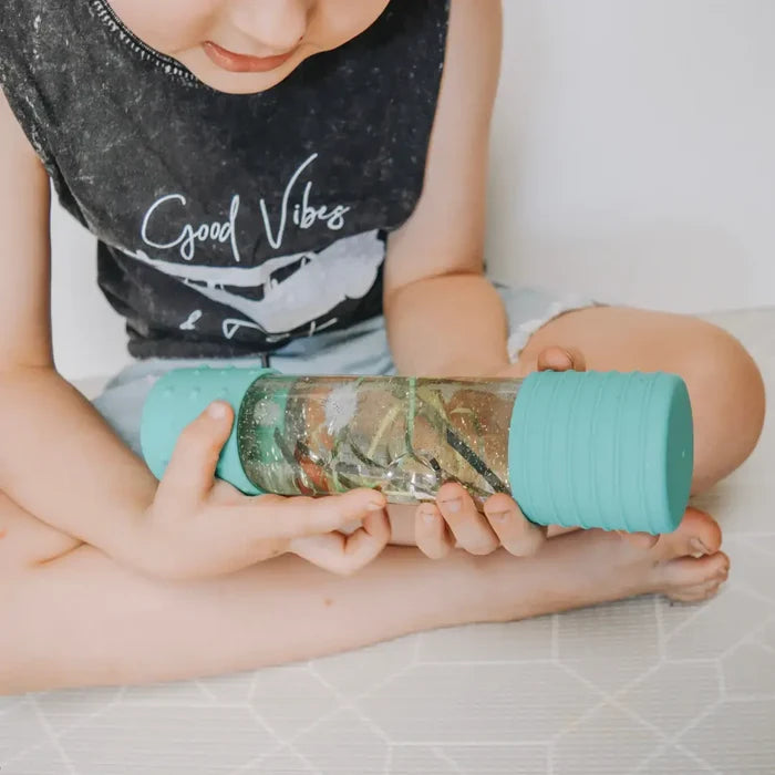 Child holding a clear sensory bottle with mint green textured caps, filled with liquid, glitter, and green items.