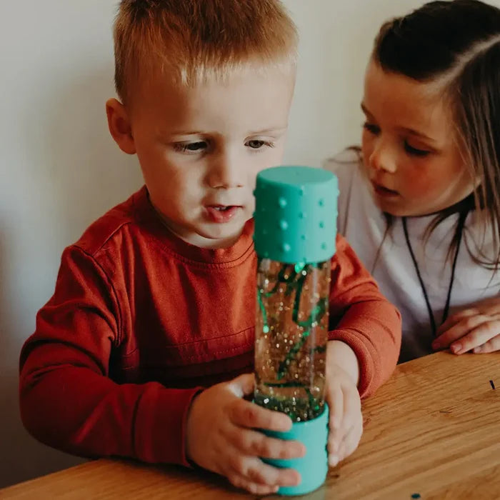 Boy holds a clear sensory bottle with mint green textured caps, filled with gold glitter and green shapes.
