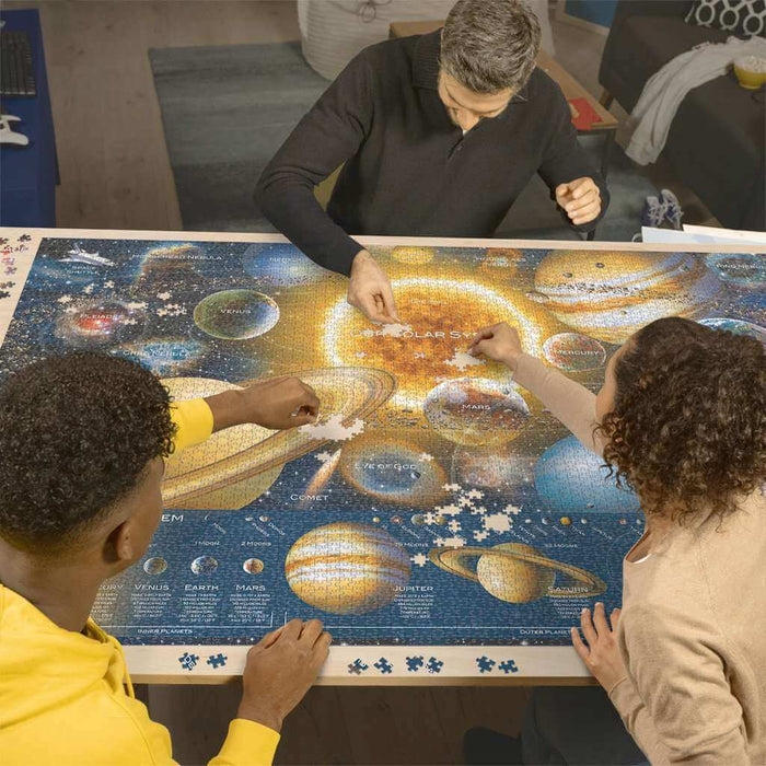 A family assembles a large solar system puzzle featuring the sun, Jupiter, Saturn, and Earth on a table.