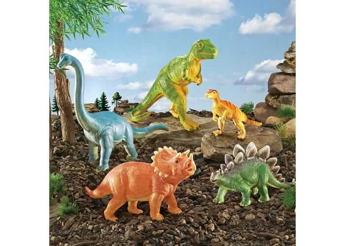 Five toy dinosaurs including a blue long-neck, green T-Rex, and orange triceratops on a rocky landscape.