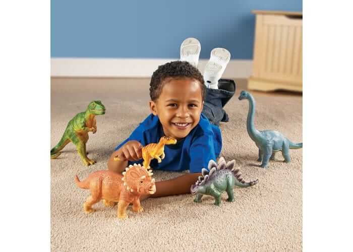 Smiling boy on a carpet with five large, colorful dinosaur toy figurines including a green T-Rex and blue Brachiosaurus.