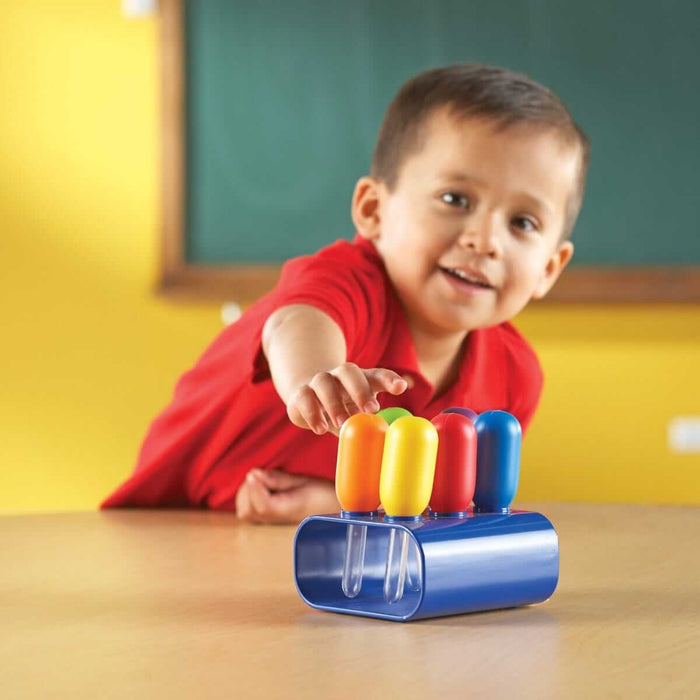 Smiling boy reaches for colorful jumbo eyedroppers with large rounded bulbs in a blue plastic stand on a wooden table.