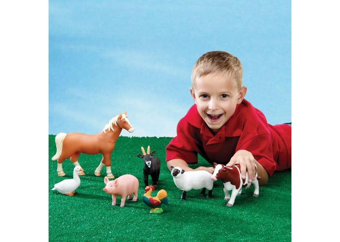 Smiling boy with jumbo plastic farm animals including a brown horse, red cow, and white sheep on green grass.
