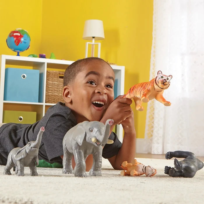 Smiling boy plays with large and small grey elephants, orange striped tigers, and a black gorilla toy on a white rug.