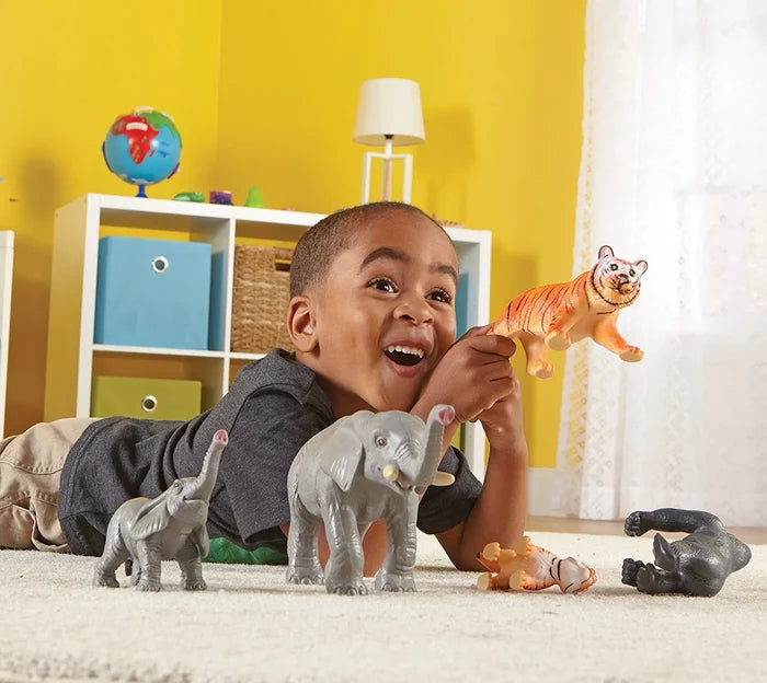 Smiling child plays with large and small grey elephant and orange tiger toy figurines on a white rug.