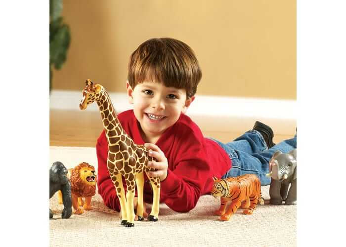 Smiling boy in a red shirt plays on a rug with large toy giraffe, lion, gorilla, tiger, and elephant figurines.