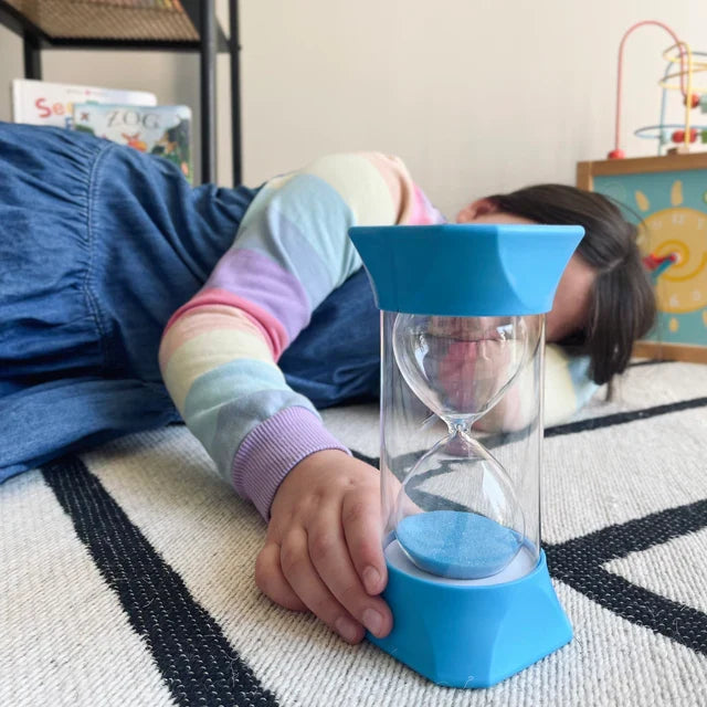 Large blue sand timer with blue sand and a clear protective shell, held by a child lying on a striped rug.