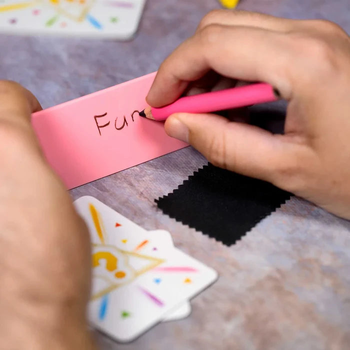 Hands writing the word Fun on a pink dry-erase board with a pink marker, next to game cards and a black eraser cloth.