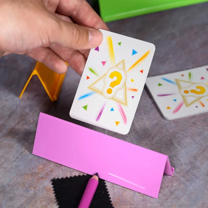 Hand placing a question mark card into a pink stand beside colorful game pieces and a pink pencil on a gray surface.