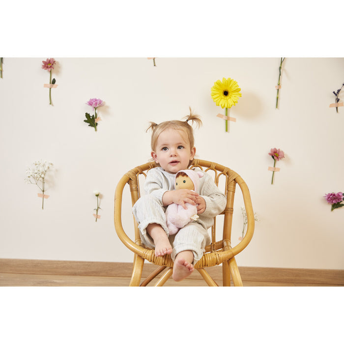 Toddler girl sits in a wicker chair holding a soft pink Kaloo baby doll against a wall decorated with taped flowers.