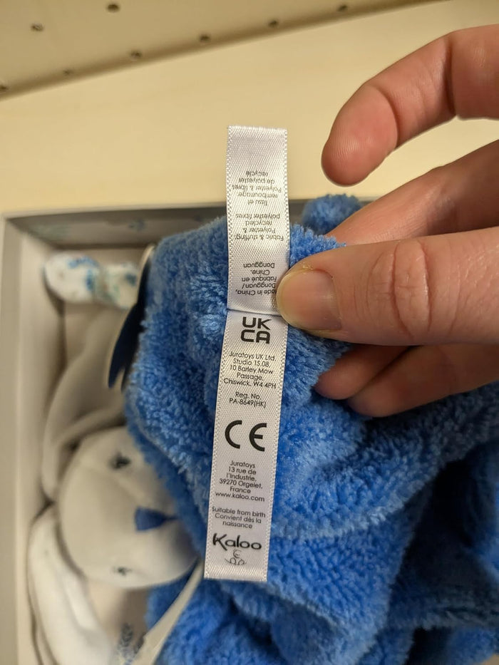 Close-up of a white tag on soft, ocean blue plush fabric, showing the Kaloo logo and safety certifications.