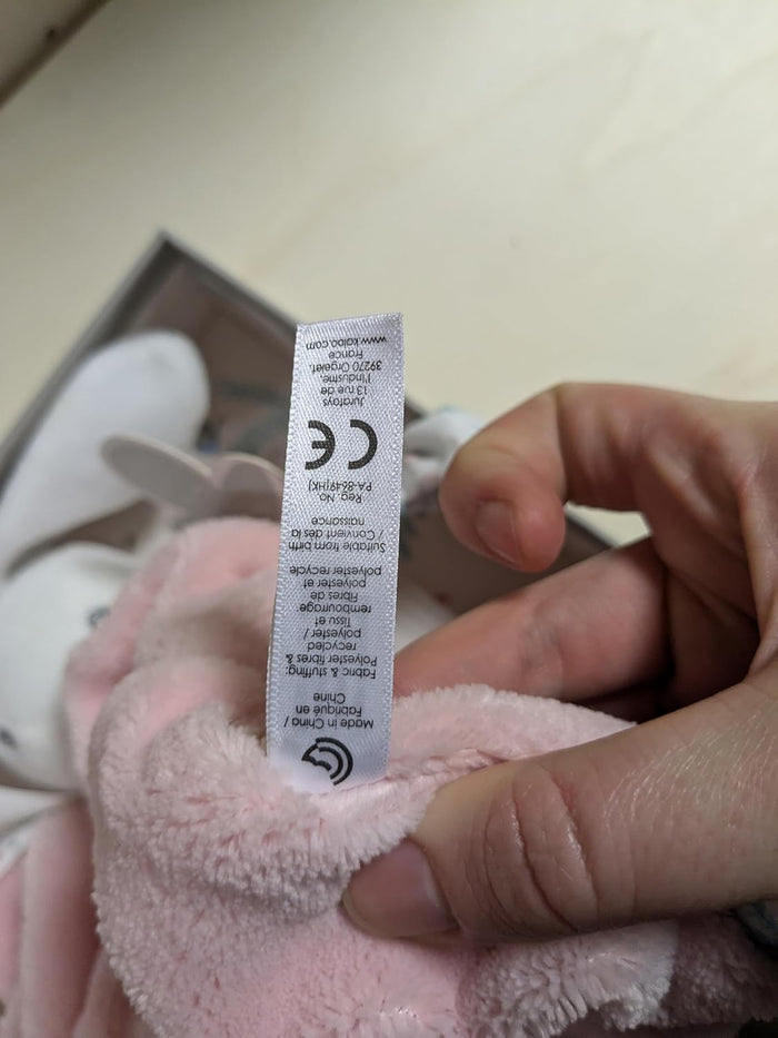 Close-up of a hand holding a white care label with a CE mark on a soft, powder pink fuzzy plush toy.