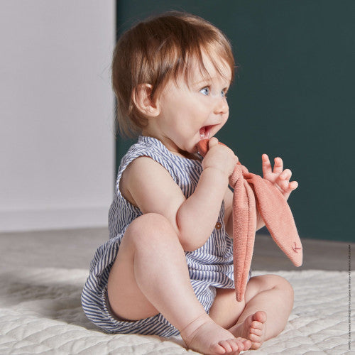 Baby sitting on a white mat chewing on a terracotta fabric rabbit ear teether.