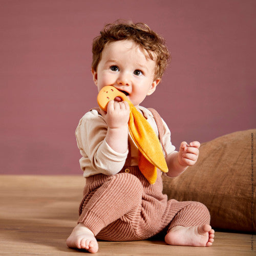 Baby biting an ochre rabbit teether with a silicone face and long fabric ears, sitting in pink knit overalls.