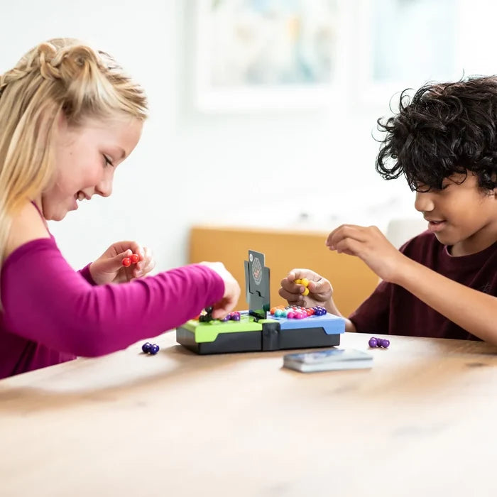 Two children play a logic puzzle game with colorful ball pieces on a green and blue board at a wooden table.