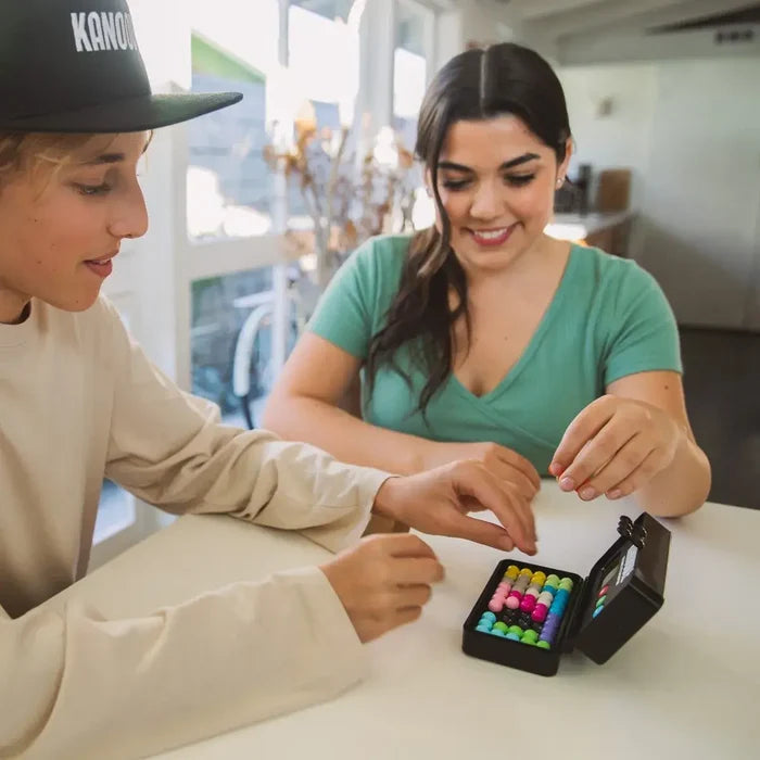 Two people solve a puzzle with colorful connected spheres in a compact black travel case on a white table.