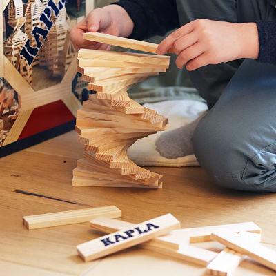 Child building a twisting spiral tower with rectangular light wood planks on a floor with Kapla blocks and box.