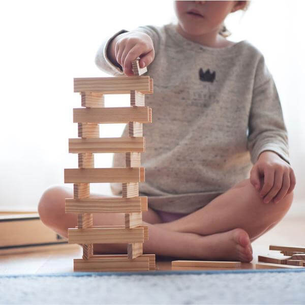 Child building a tall tower with rectangular natural wood planks on a light-colored floor.