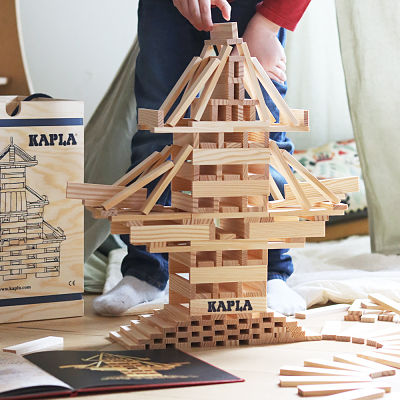 Child builds a wooden plank tower next to the open Kapla Art Book Red with a black and gold cover.